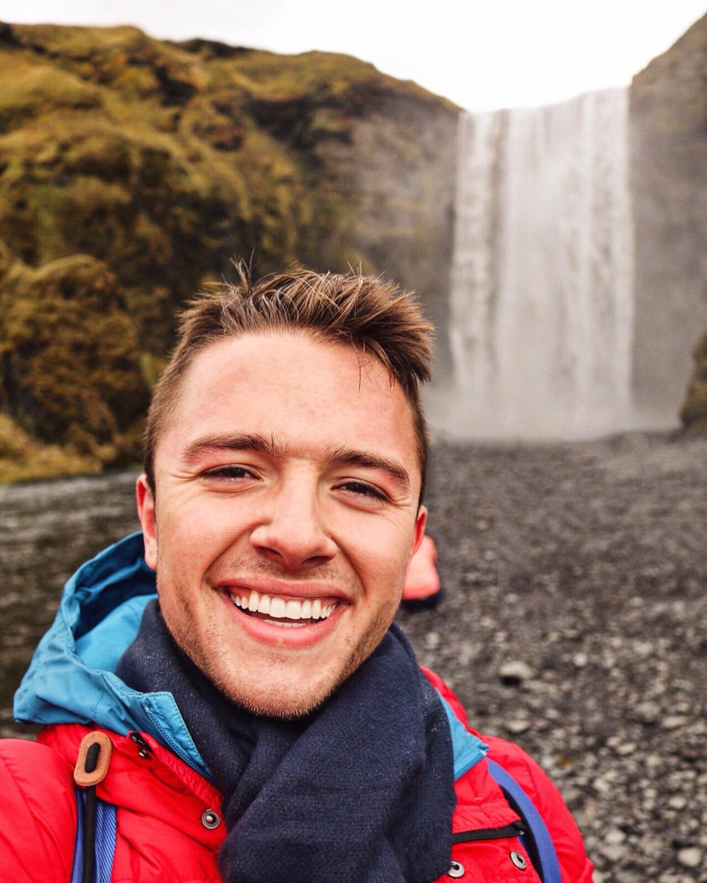 a picture of Nick, a 24-year-old man with short brown hair, brown eyes, and a bright smile poses for a selfie in the foreground with a waterfall in the background - Nick is wearing a red winter coat over a blue rain coat, and a cotton navy-colored scarf